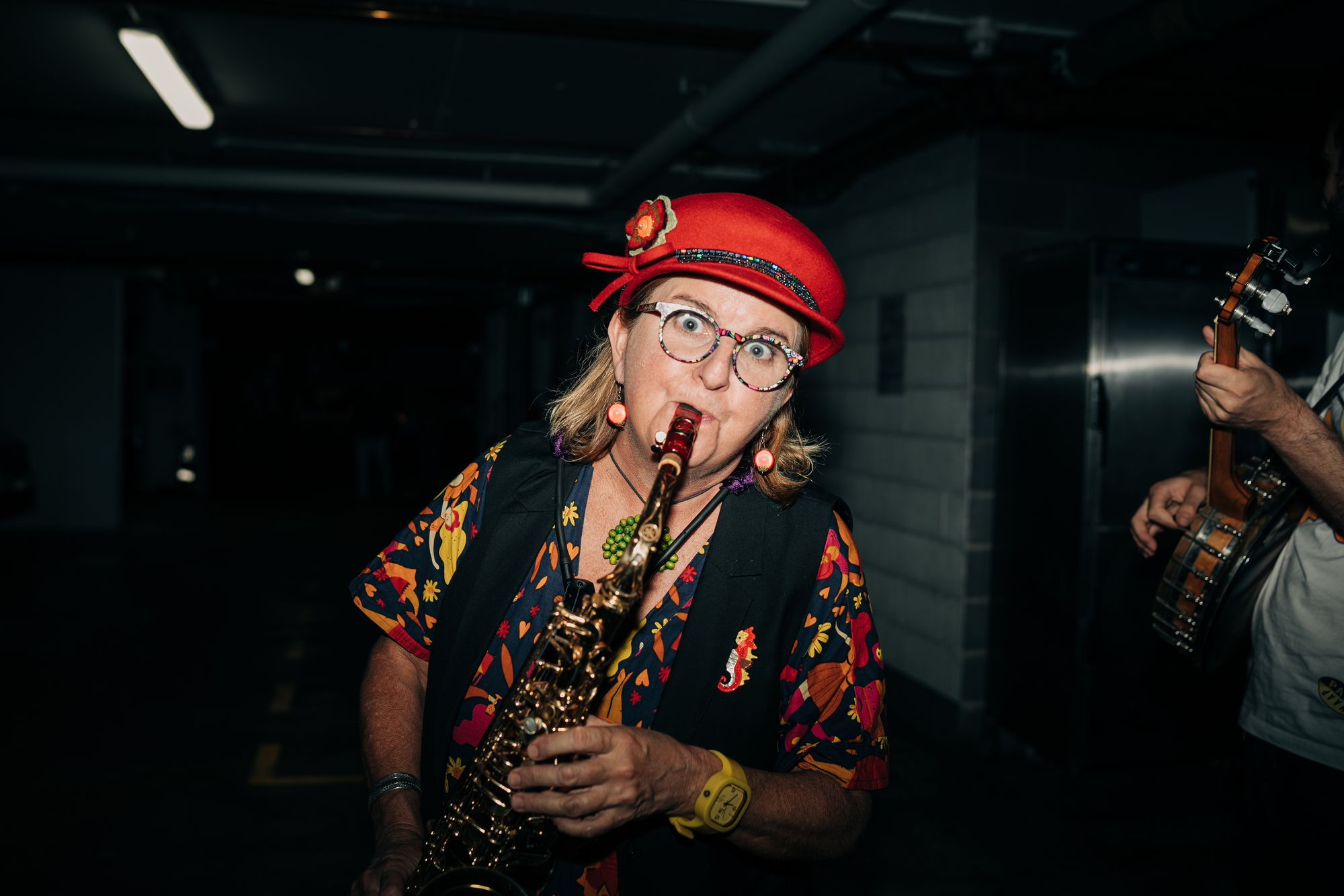 A woman in a red hat and colorful outfit plays a saxophone passionately in a dimly lit room.