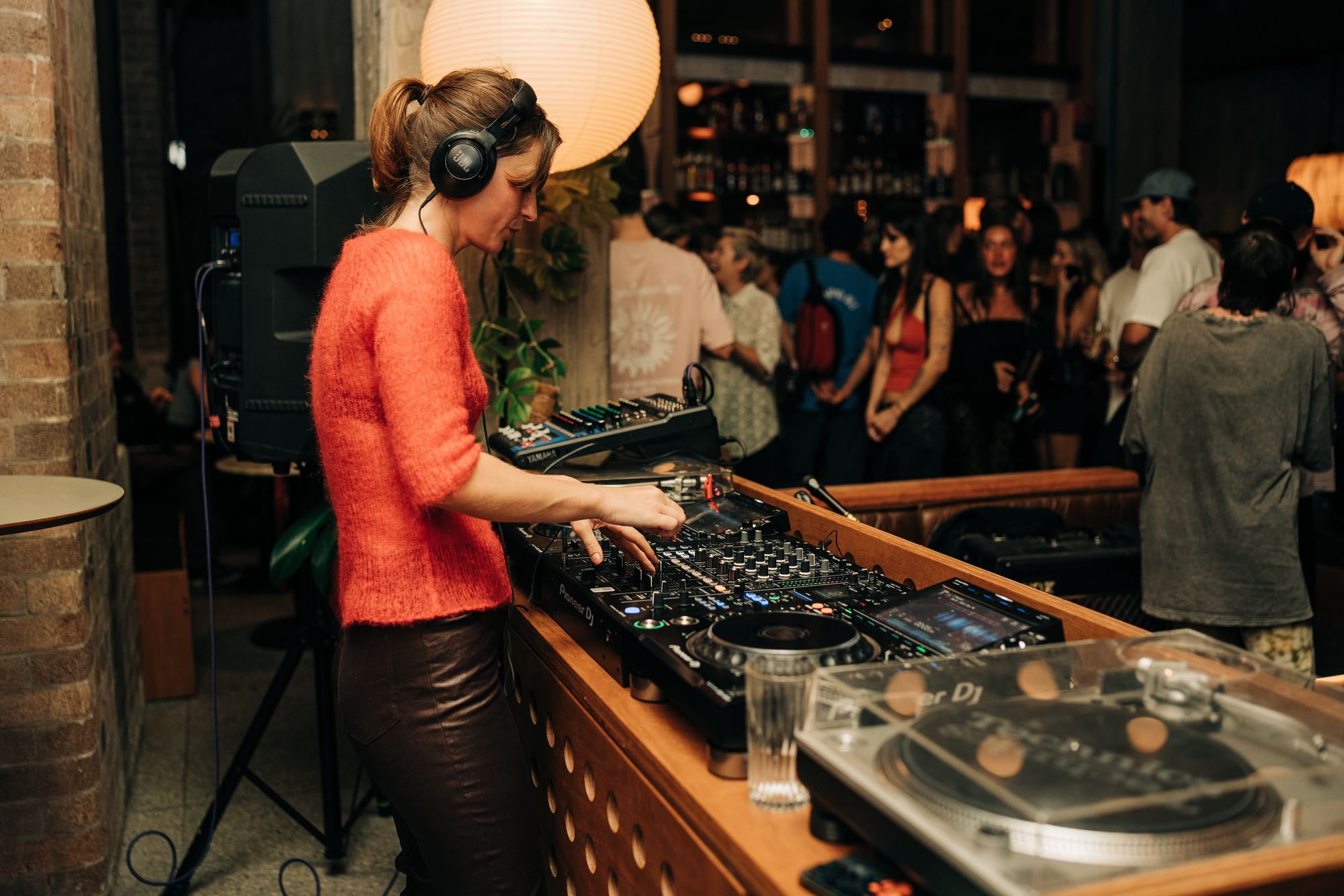 DJ wearing headphones mixes music on a turntable in a lively bar with a crowd in the background.