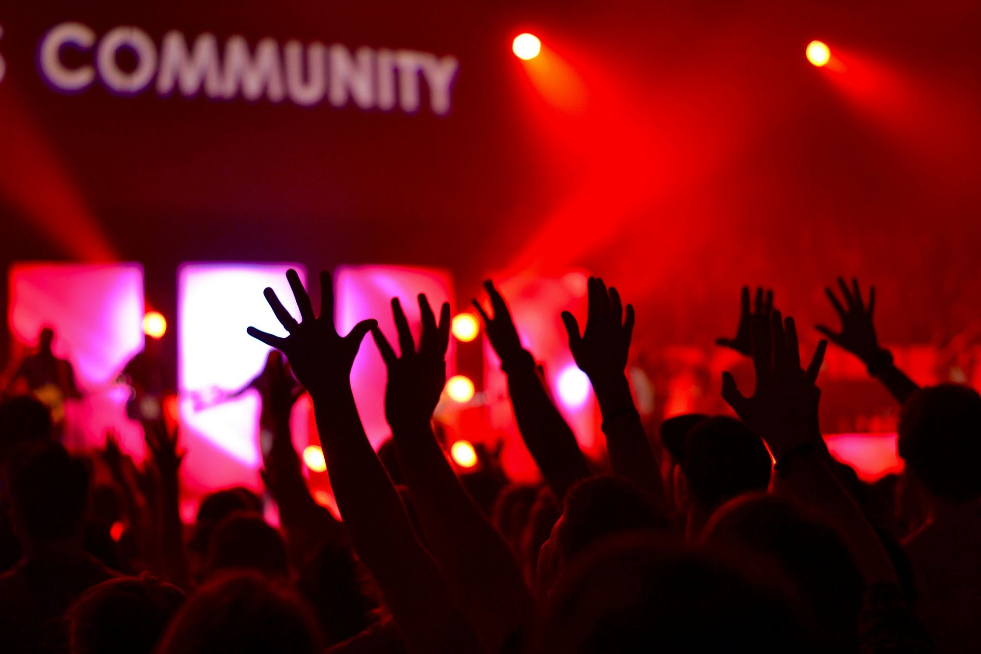 Silhouetted crowd with raised hands at an event, under red lighting. The word "COMMUNITY" is partially visible on a screen in the background.
