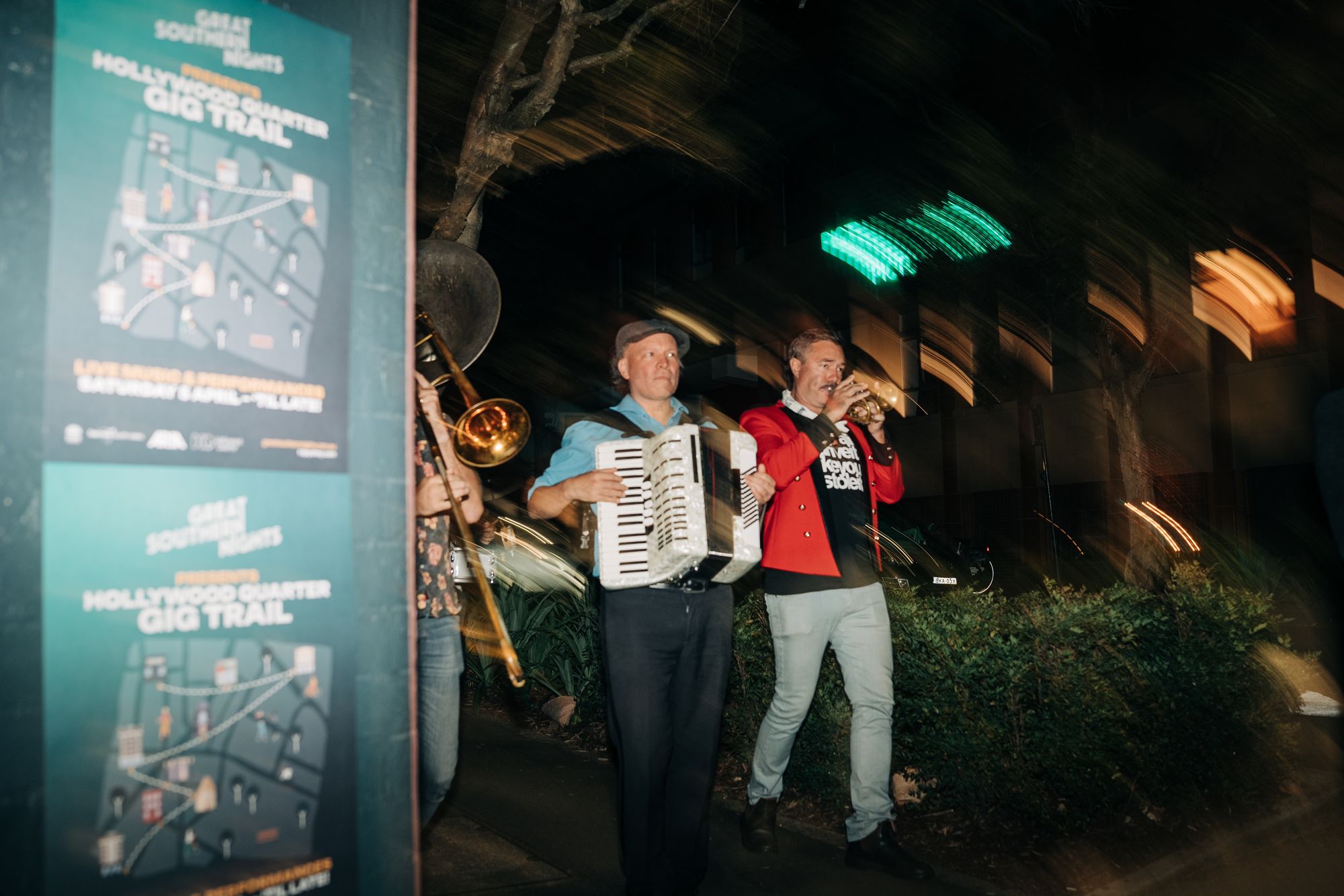 Musicians play accordion and trumpet while walking outdoors at night, with blurred lights and event posters in the background.