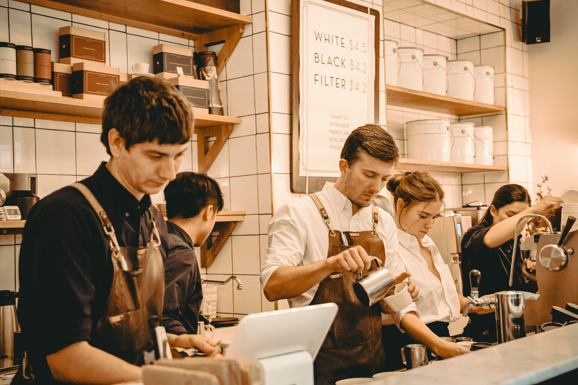 Baristas wearing aprons prepare drinks behind a coffee shop counter, with shelves of supplies and a menu board in the background.