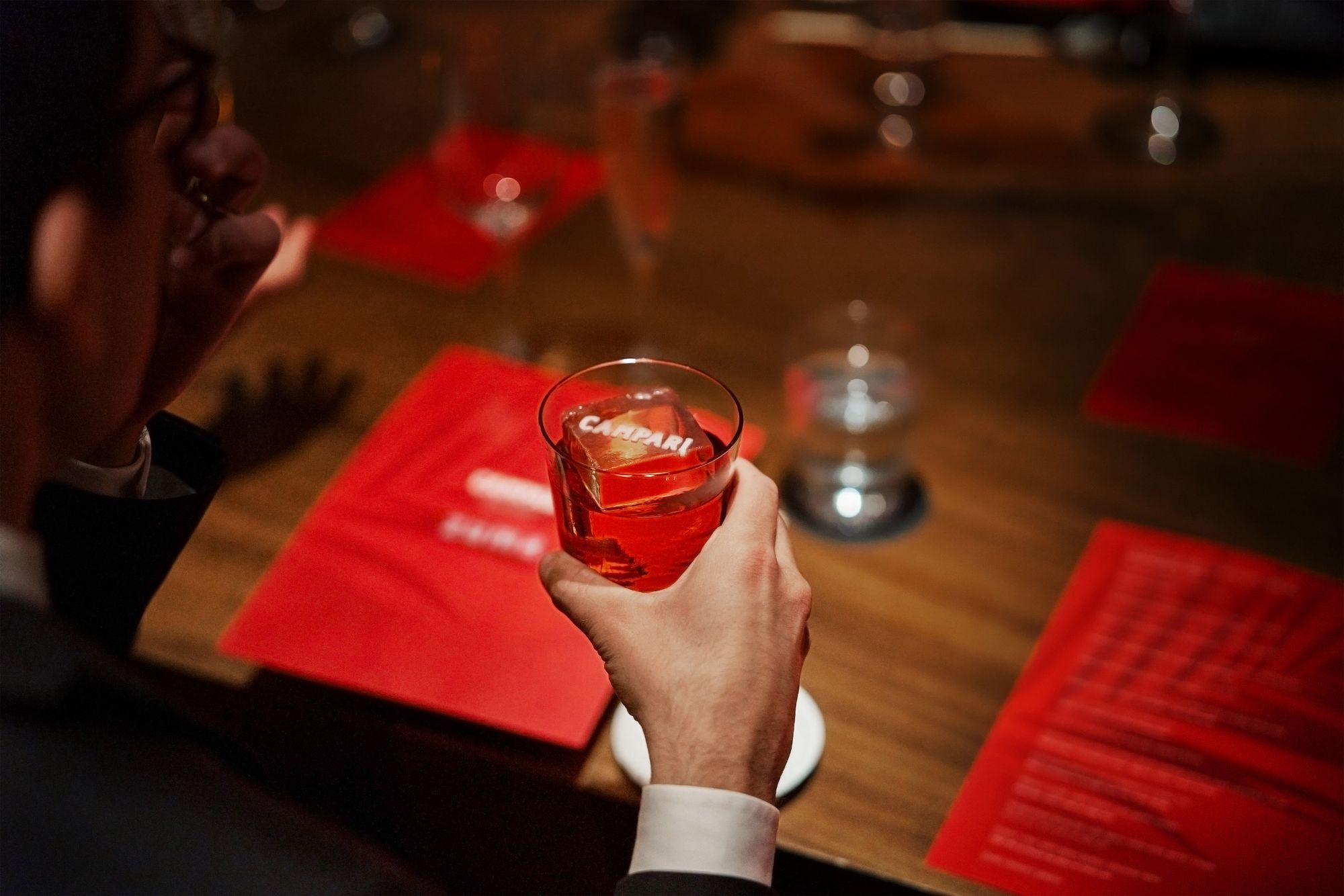Person holding a glass of red cocktail at a dimly lit table with red menus and a champagne flute.