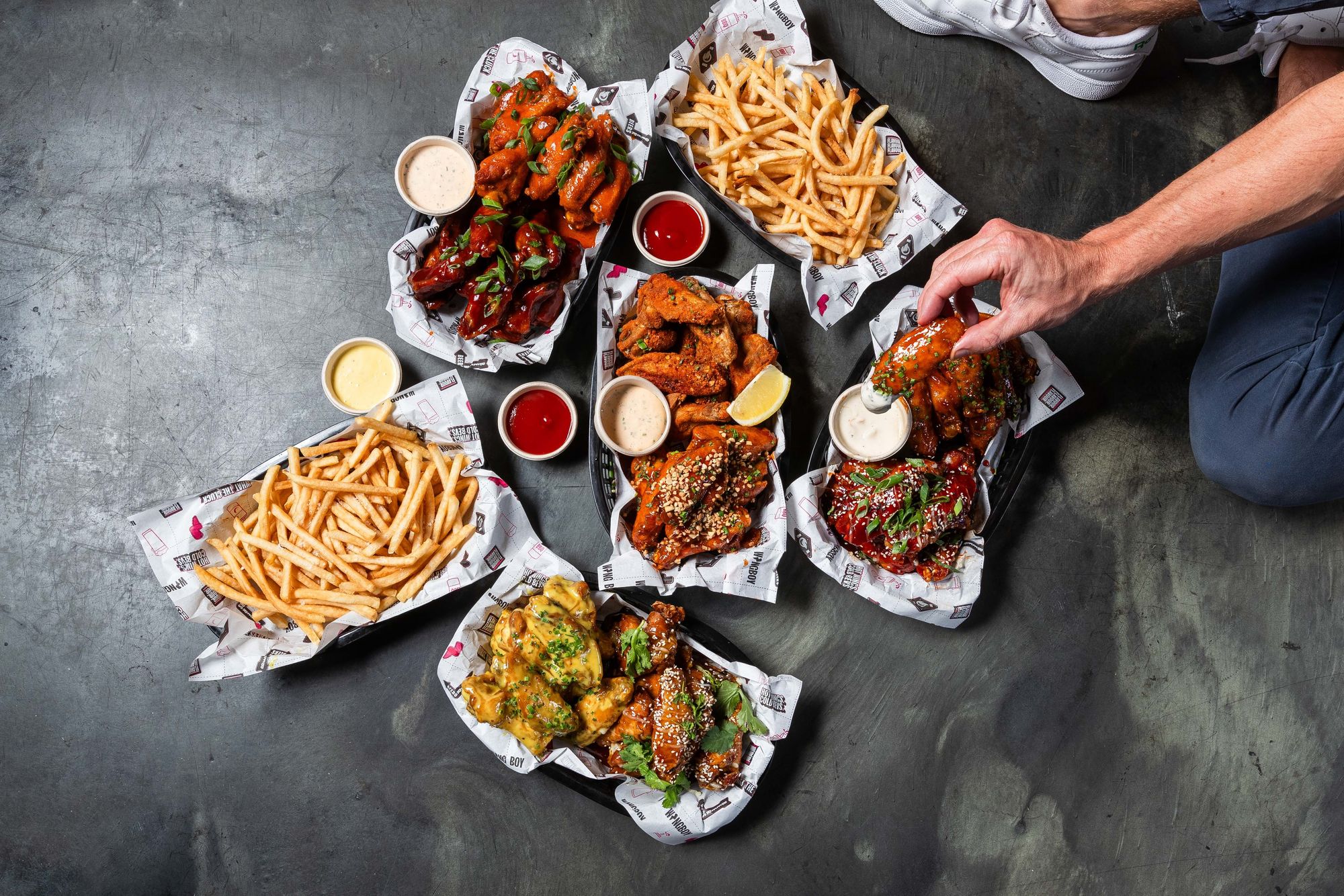 A variety of seasoned chicken wings and fries in baskets with dipping sauces. A hand reaches for a wing.