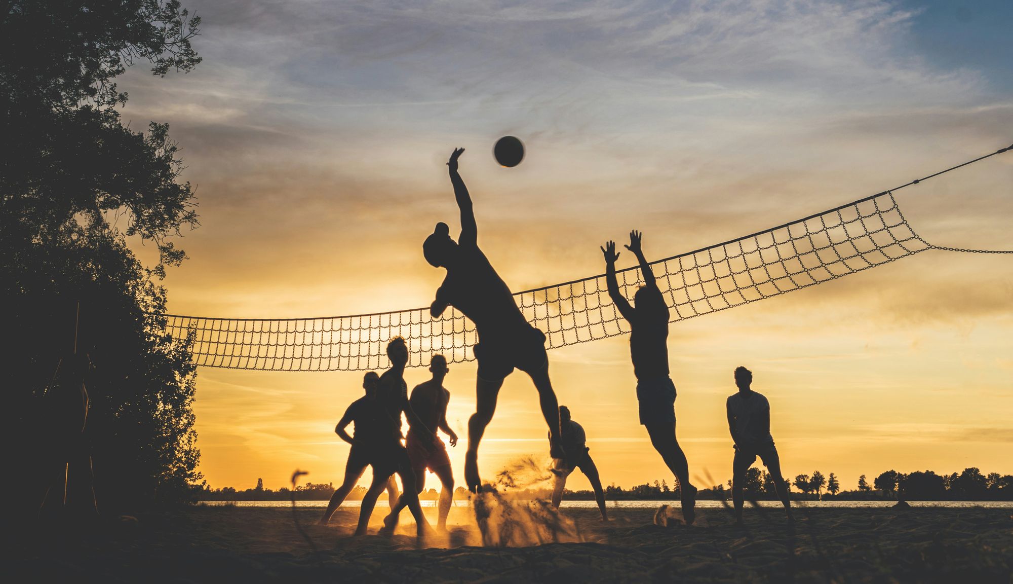 Silhouetted group playing beach volleyball at sunset, with one player leaping to hit the ball over the net.