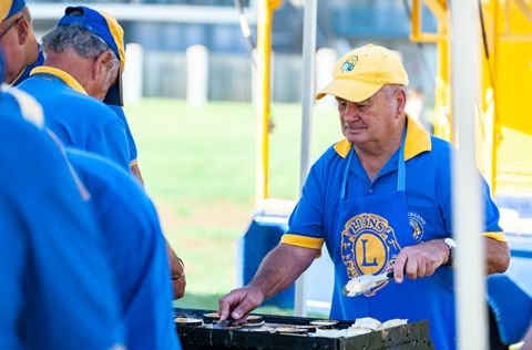 A man in a Lions Club shirt grills food at an outdoor event, using tongs and wearing a yellow cap.