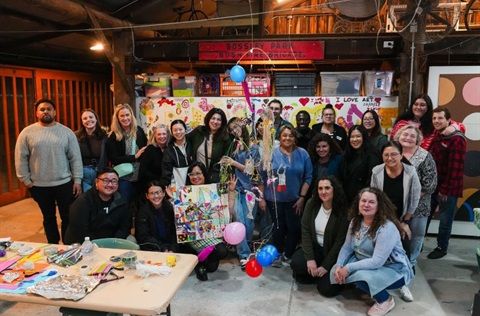 A group of people poses with art supplies and colourful decorations in a lively indoor setting. Some hold a creative display board.