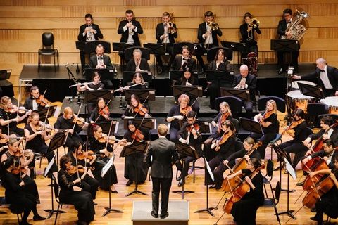 Orchestra performing on a stage, conductor in front. Musicians play various instruments like violins, cellos, and brass.