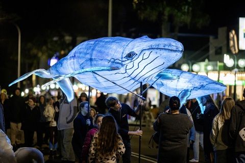 People carrying illuminated whale-shaped lanterns at a night parade, surrounded by onlookers on a city street.