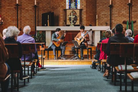 Two musicians play guitars on a stage in a church, with a seated audience watching. The background features brick walls and a stained glass window.