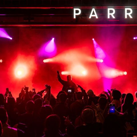 Concert crowd in dim lighting with red and purple spotlights. A performer is silhouetted on stage holding a microphone.