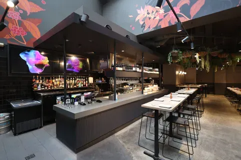 Modern restaurant interior with a long bar counter, high stools, contemporary lighting, and artistic wall designs featuring floral patterns.