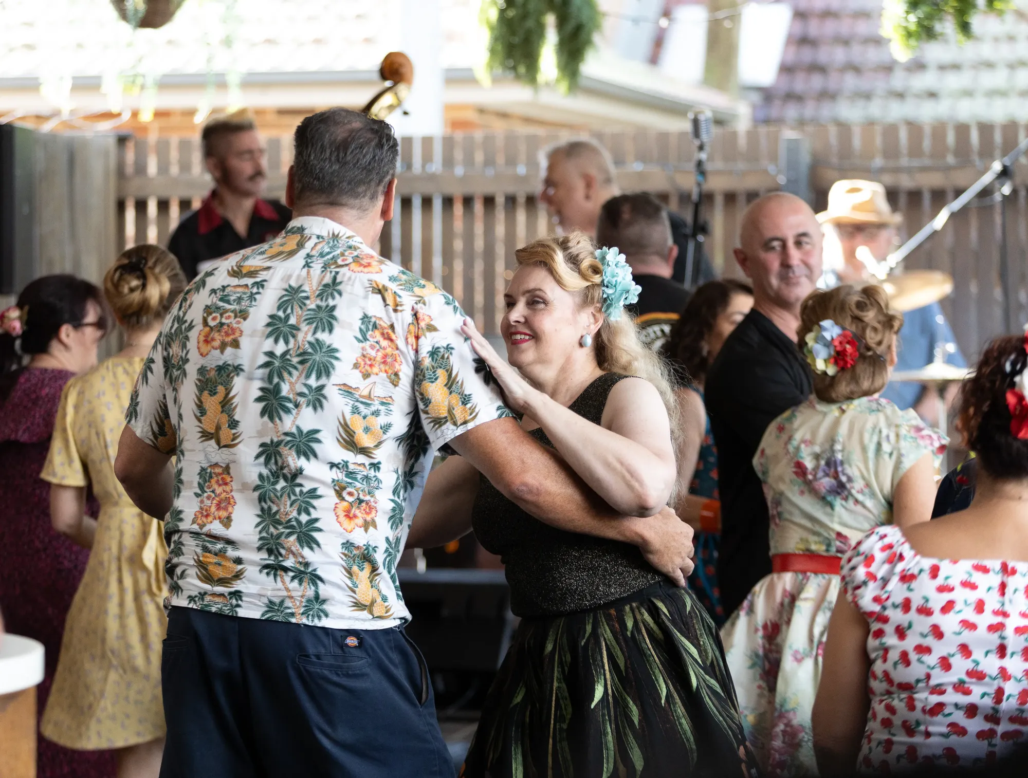 People in vintage clothing dancing joyfully at an outdoor event, with musicians playing in the background and colorful decorations.