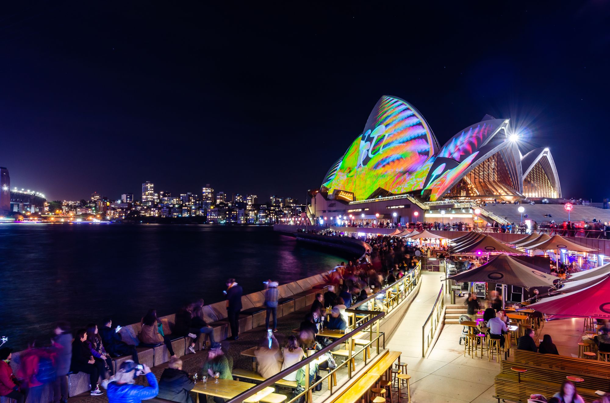 Sydney Opera House lit up with colorful projections during a nighttime event, with waterfront crowds enjoying the vibrant scene.