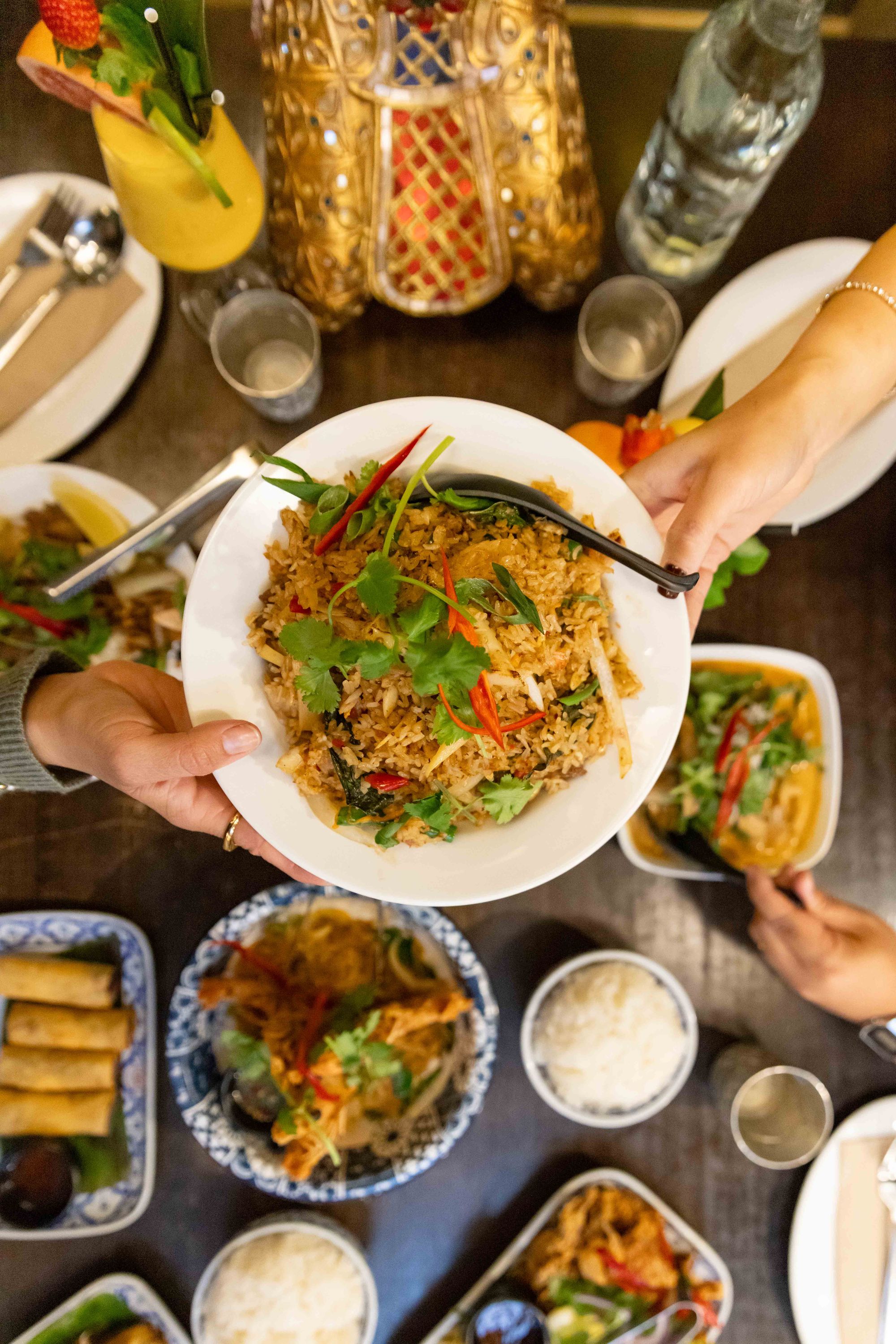 Hands passing a plate of fried rice over a table with various Asian dishes, including spring rolls, noodles, and rice, surrounded by drinks and decor.