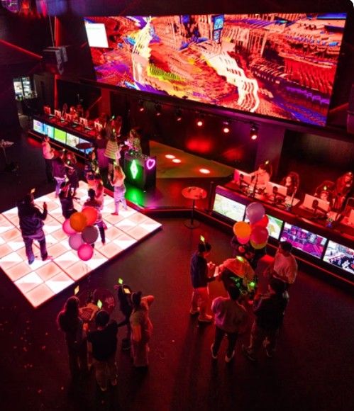 A vibrant nightclub with people dancing on an illuminated floor, colorful balloons, and a large digital display overhead.