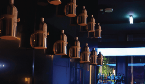 Hanging large metallic jugs in a dimly lit modern bar, with a glimpse of bottles on shelves in the background.