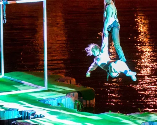Performers suspended over water in a night show, with one artist holding another by the arm, illuminated by colorful lights.