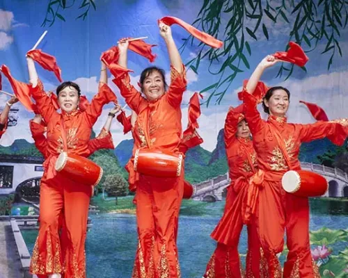 Women in red costumes perform a traditional dance with drums and red ribbons against a scenic backdrop of water and greenery.