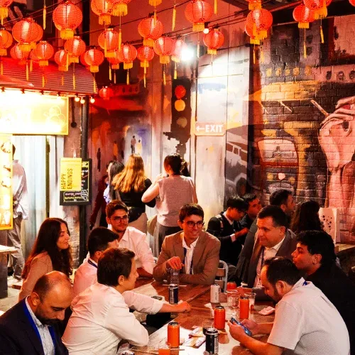 People sit at a dimly lit restaurant with red lanterns hanging overhead and vibrant artwork on brick walls, enjoying conversation and drinks.