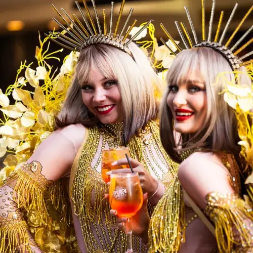 Two smiling women in ornate gold costumes and spiked crowns, holding drinks, surrounded by yellow decorative elements.