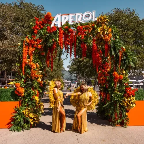 Two people in gold outfits pose under a vibrant floral arch with an "Aperol" sign, set outdoors with trees in the background.