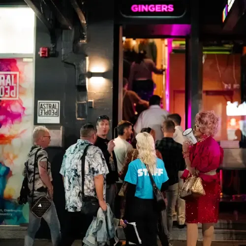 People gather outside a nightlife venue called "Gingers" under vibrant lighting. A staff member and individuals in diverse attire are visible.