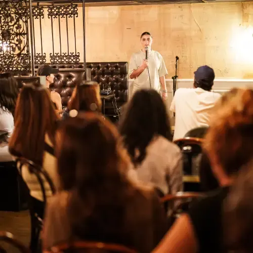 Person standing on a small stage speaking into a microphone, facing an attentive seated audience in a dimly lit room.