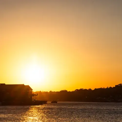 A tranquil sunset over Sydney Harbour, with a silhouetted building on the left, casting a golden reflection on the water, and an orange-hued sky.