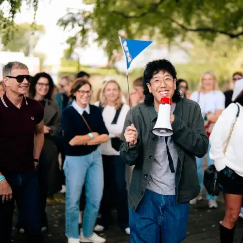 A person holding a megaphone and small flag speaks to a smiling crowd outdoors.