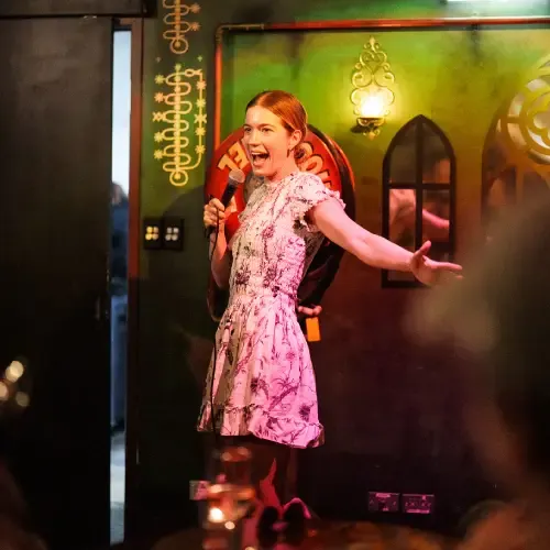 A woman in a floral dress, holding a microphone, enthusiastically speaks to an audience in a dimly lit room with decorative walls.