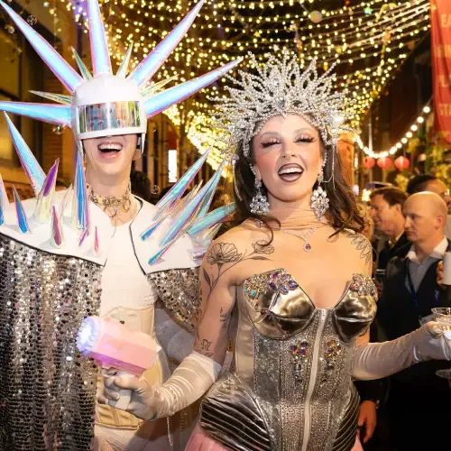 Two people in extravagant, futuristic costumes with spikes and glittering headpieces, smiling under festive lights in a crowded street.