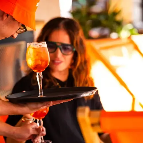 A waiter serves a refreshing orange cocktail to a woman with long hair and sunglasses, sitting outdoors in bright, warm lighting.