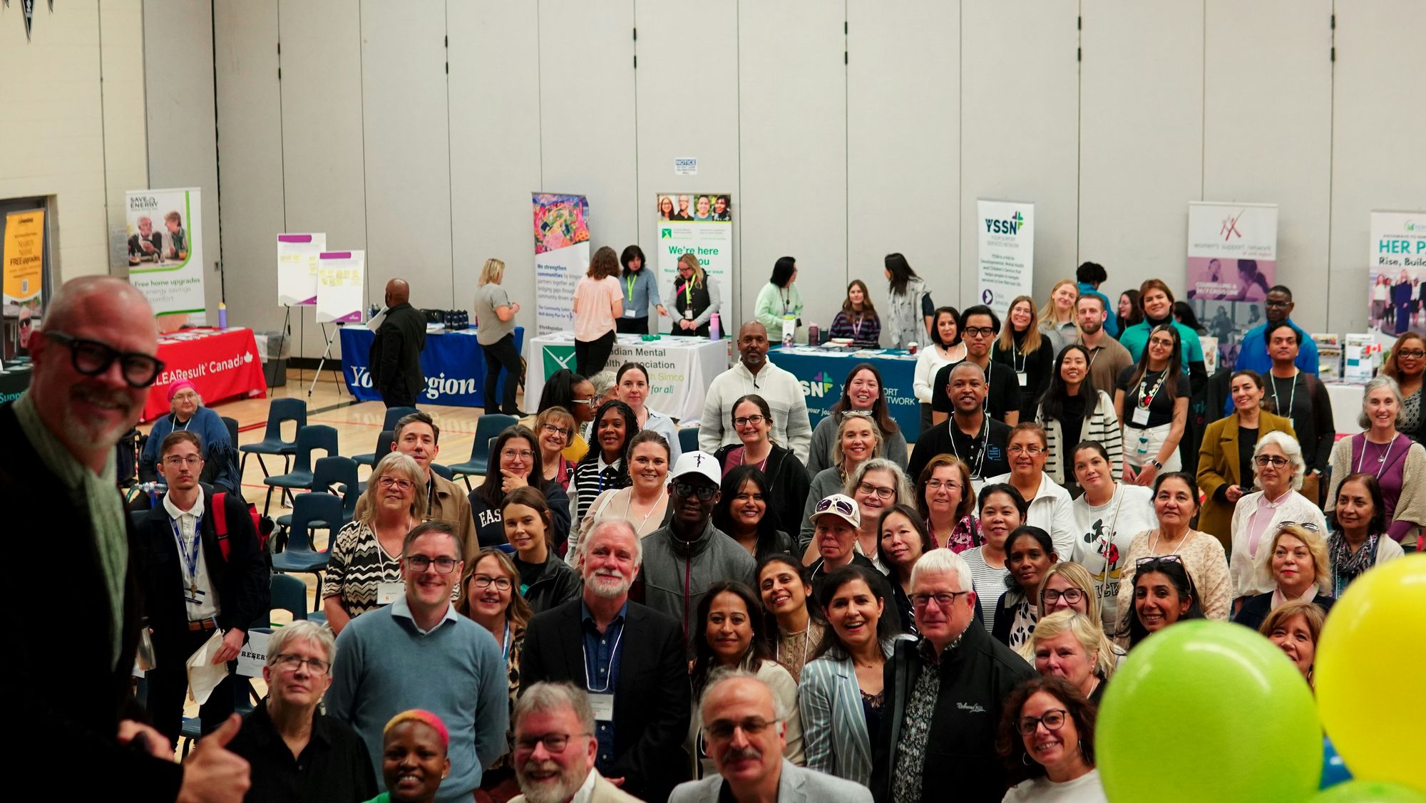 A large diverse group of people smiling at a community event with informational booths and banners in the background.