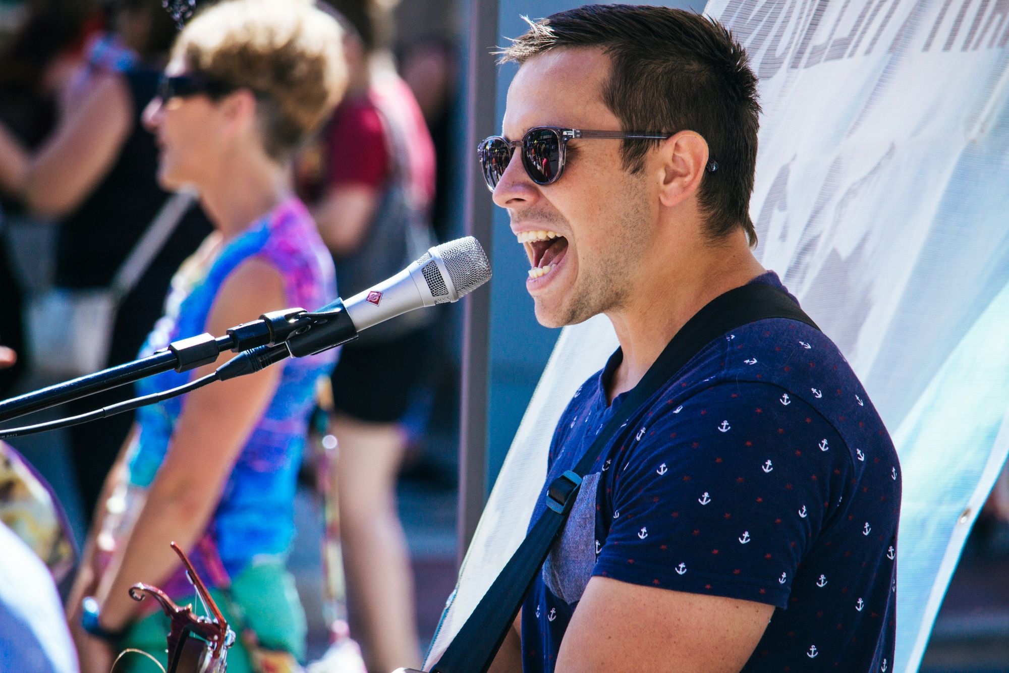 Man singing into a microphone at an outdoor event, wearing sunglasses and a patterned shirt. People in colorful clothing are in the background.