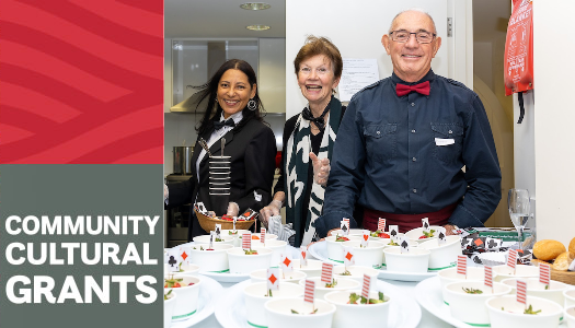 Three smiling people stand behind a table of prepared dishes at a "Community Cultural Grants" event.