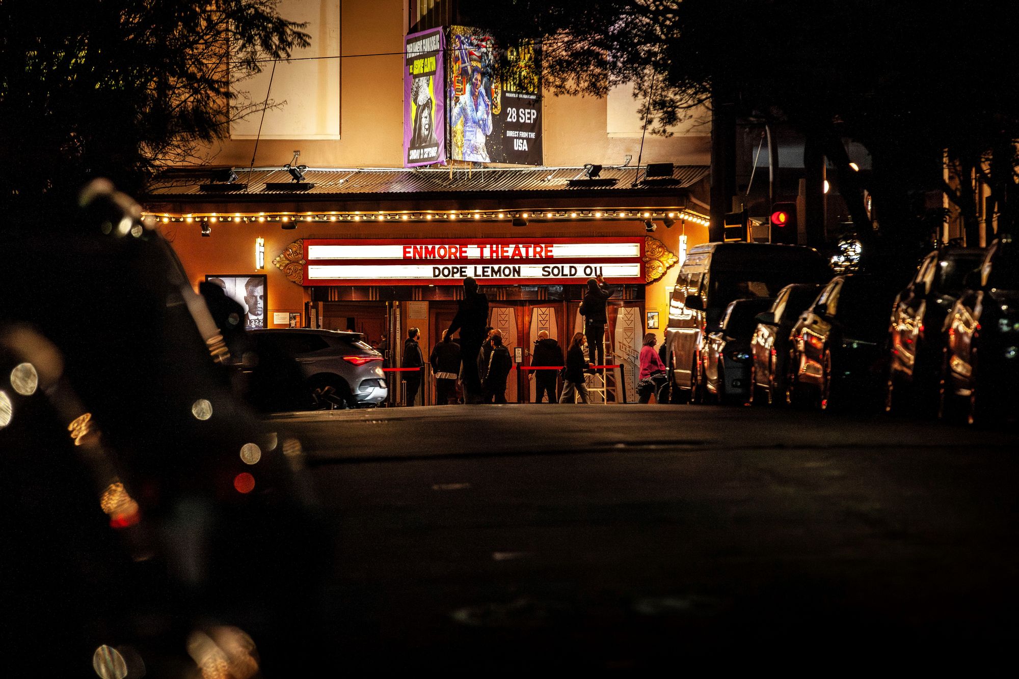 A dimly lit street with cars parked along the sides, leading to a warmly lit theater with a marquee reading "Dope Lemon Sold Out."