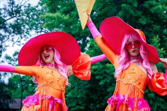 Two performers in bright pink hats and costumes joyfully pose outdoors, each holding a large ice cream cone prop, with trees in the background.