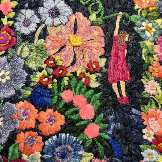 Colorful floral embroidery with a small figure in a pink dress hanging from a green vine among vibrant flowers on a dark background.