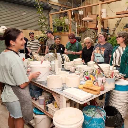 A group of people in a pottery studio gather around a table with pottery tools and materials, while a woman in an apron leads the session.