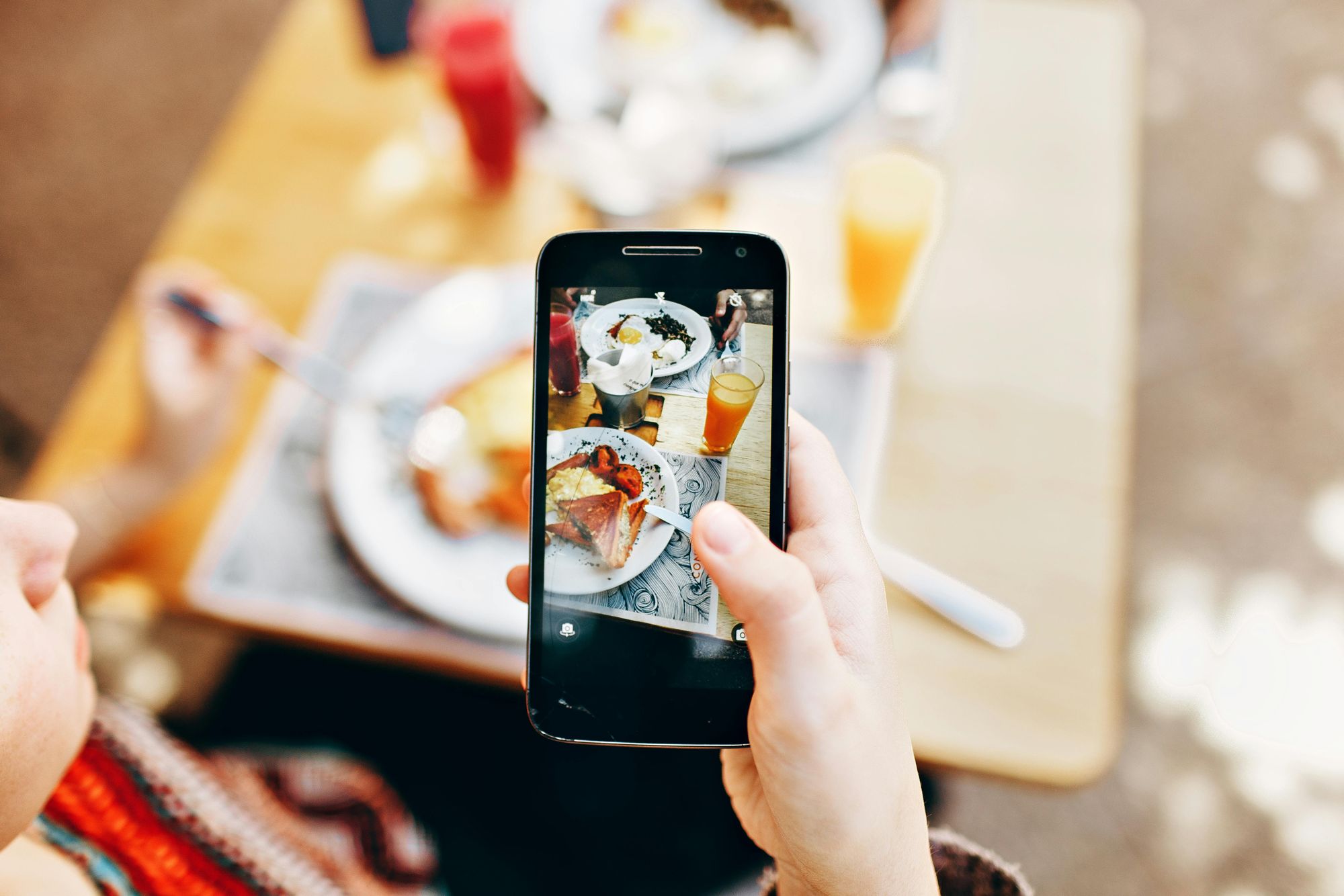 Person taking a photo of a meal with a smartphone, featuring plates of food and glasses of juice on a wooden table.