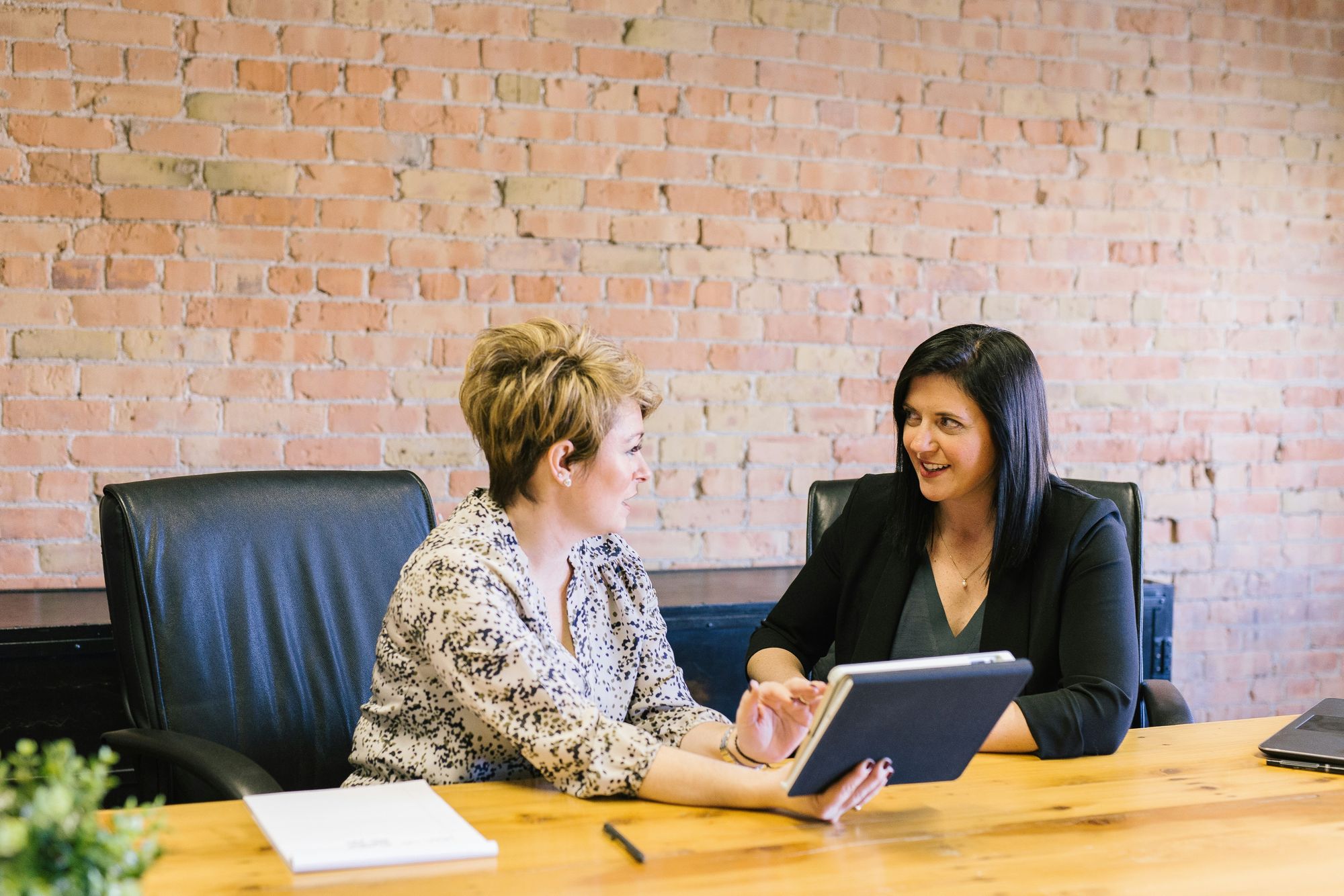 Two women sitting at a wooden table in an office, discussing something on a tablet. Brick wall background and office chairs are visible.