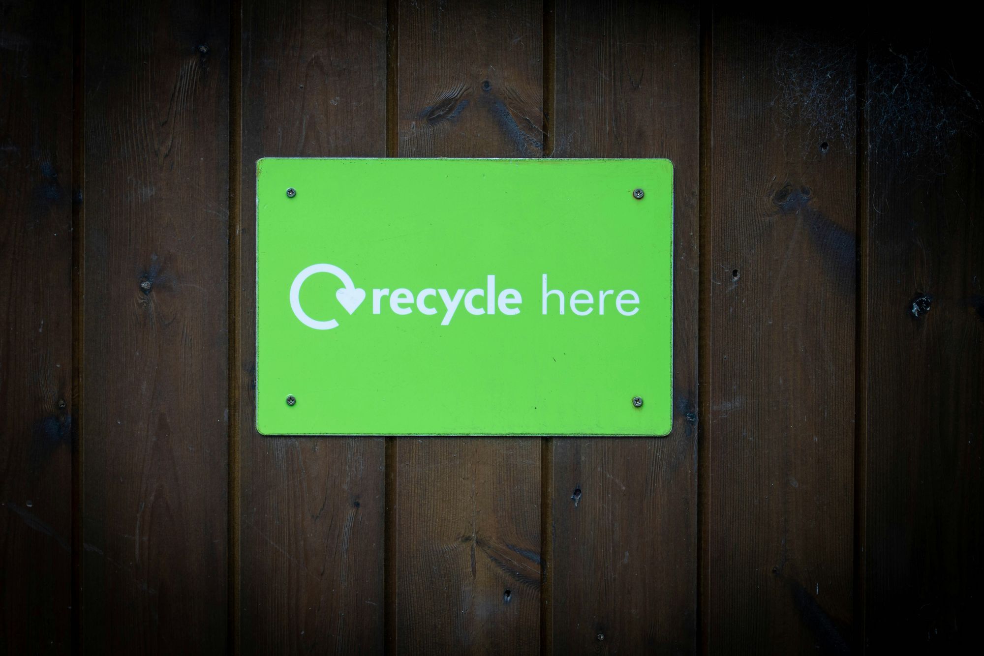 Bright green sign reading 'recycle here' with circular arrow heart icon, mounted on dark wooden plank wall.