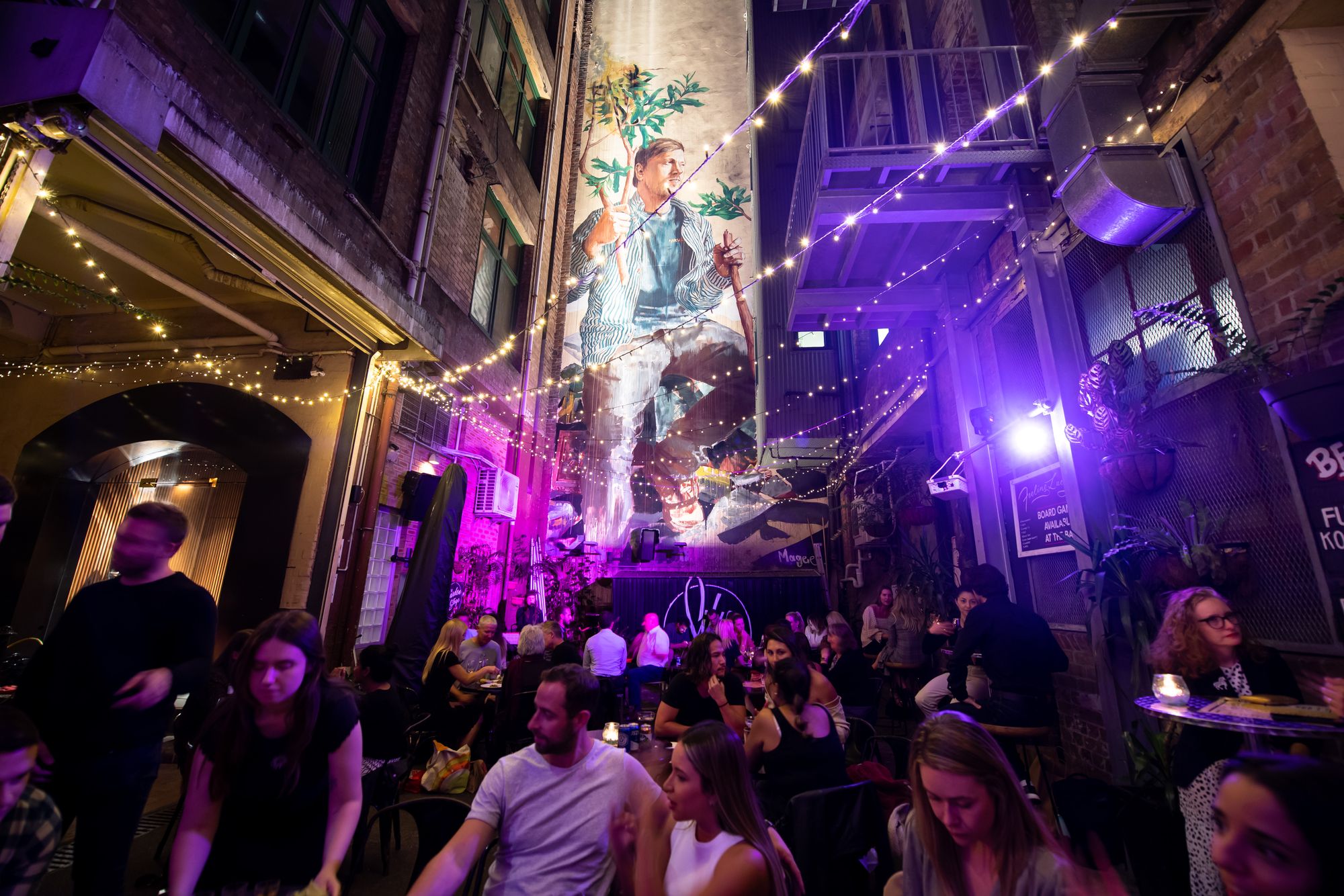 A lively alleyway bar at night with string lights, a large mural, and people socializing at tables under purple lighting.