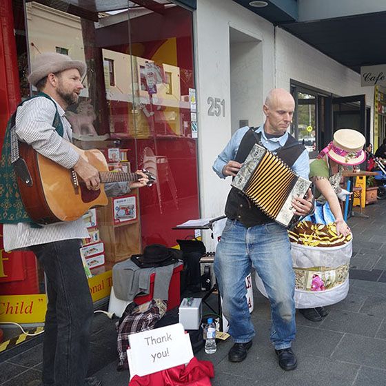 Street musicians perform with guitar and accordion, surrounded by colorful hats and a "Thank you!" sign in front of a storefront.