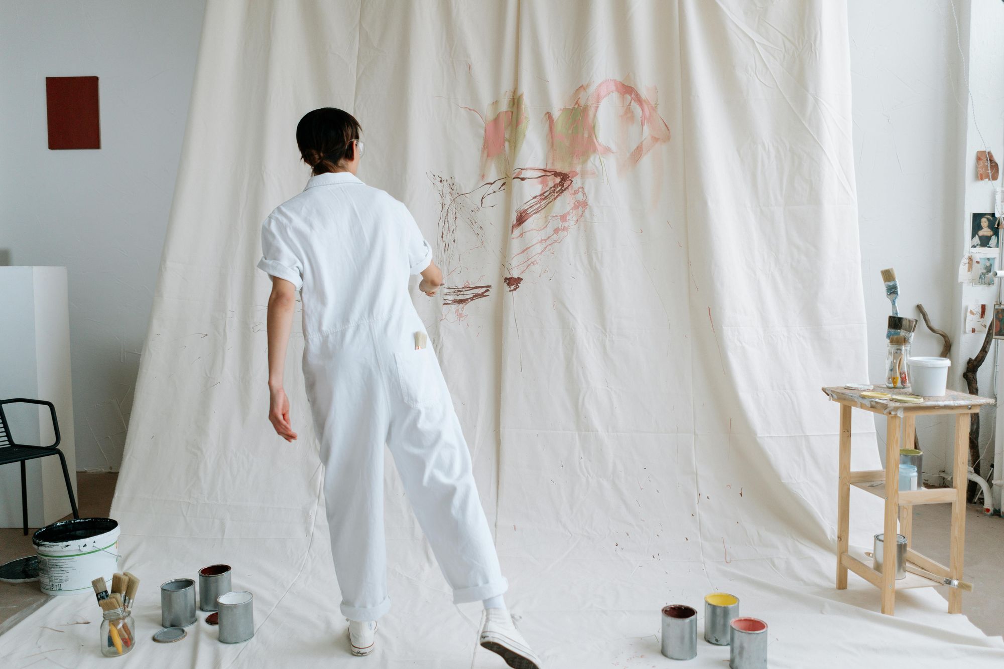 Artist in white coveralls painting abstract art on a large canvas draped on a wall, with paint cans and brushes on the floor.