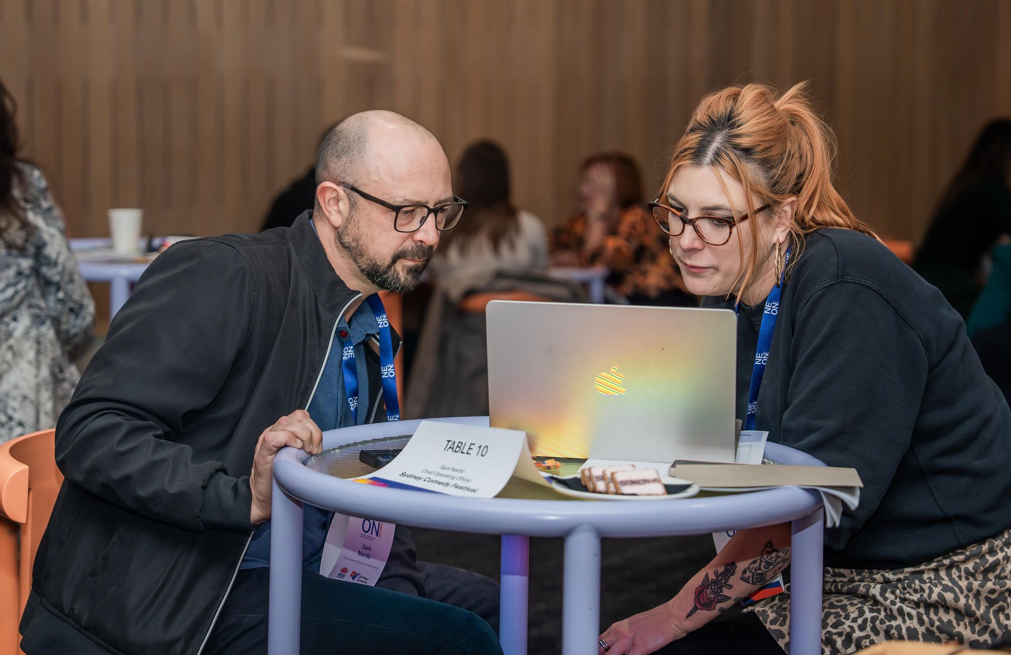 Two people seated at a round table, focused on a laptop. A sign reads "Table 10." They appear to be discussing something intently.