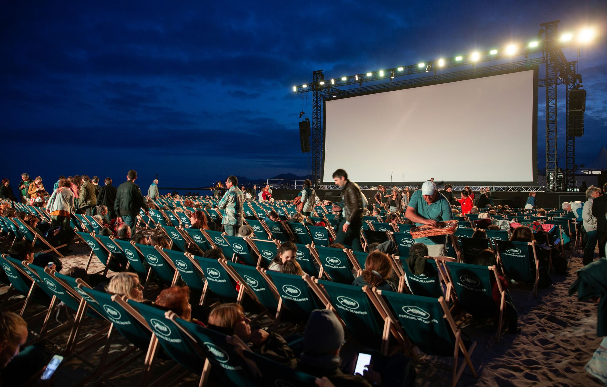 Outdoor cinema on a beach at dusk, with rows of deck chairs filled with people facing a large screen.