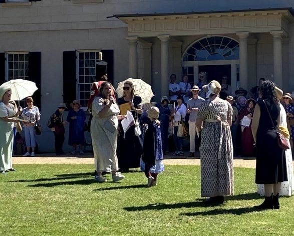 People wearing period costumes gather on a lawn in front of a historic building, some holding parasols, during a reenactment event.