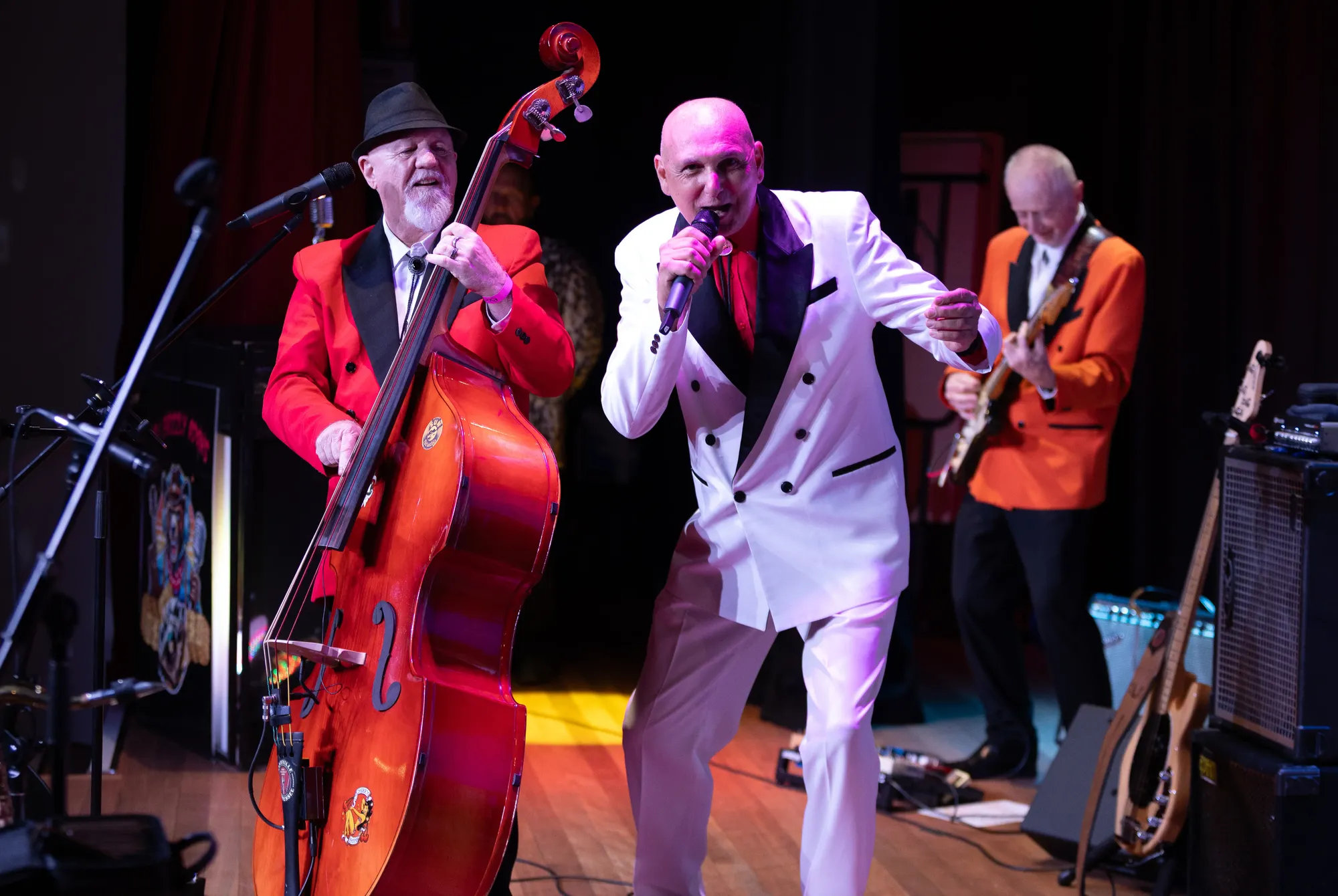 A lively band performs on stage with a double bassist in red, a singer in white, and a guitarist in orange, under colorful stage lights.