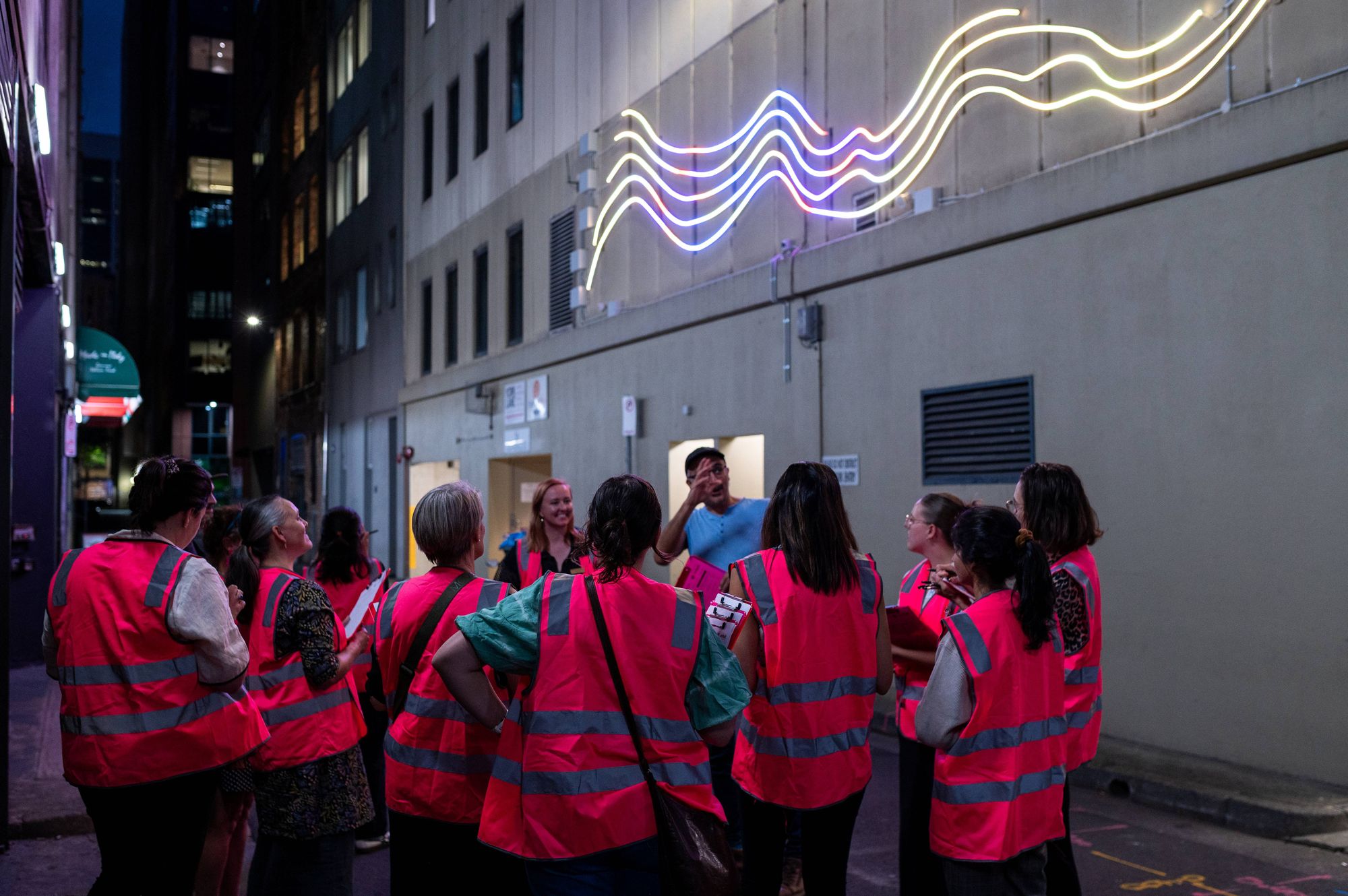 Women in pink high vis vests and clipboards participate in a perceptions of safety co-design walkshop at night with Cobalt Engamgement giving feedback on the lighting public artwork, 'Lightstream' in York Lane, Wynyard for YCK Laneways.